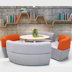 Conjunto de asientos modulares en tonos grises y naranjas alrededor de una mesa circular en un entorno de oficina.